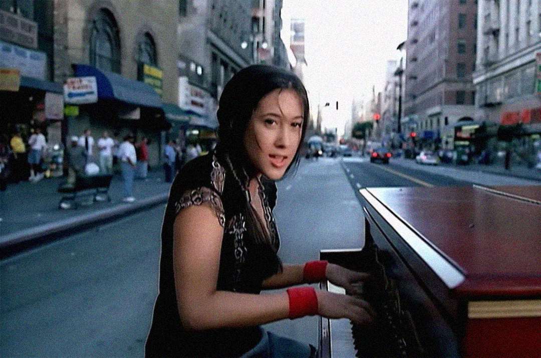A woman plays a piano on a city street, surrounded by tall buildings and blurred pedestrians, while looking toward the camera.