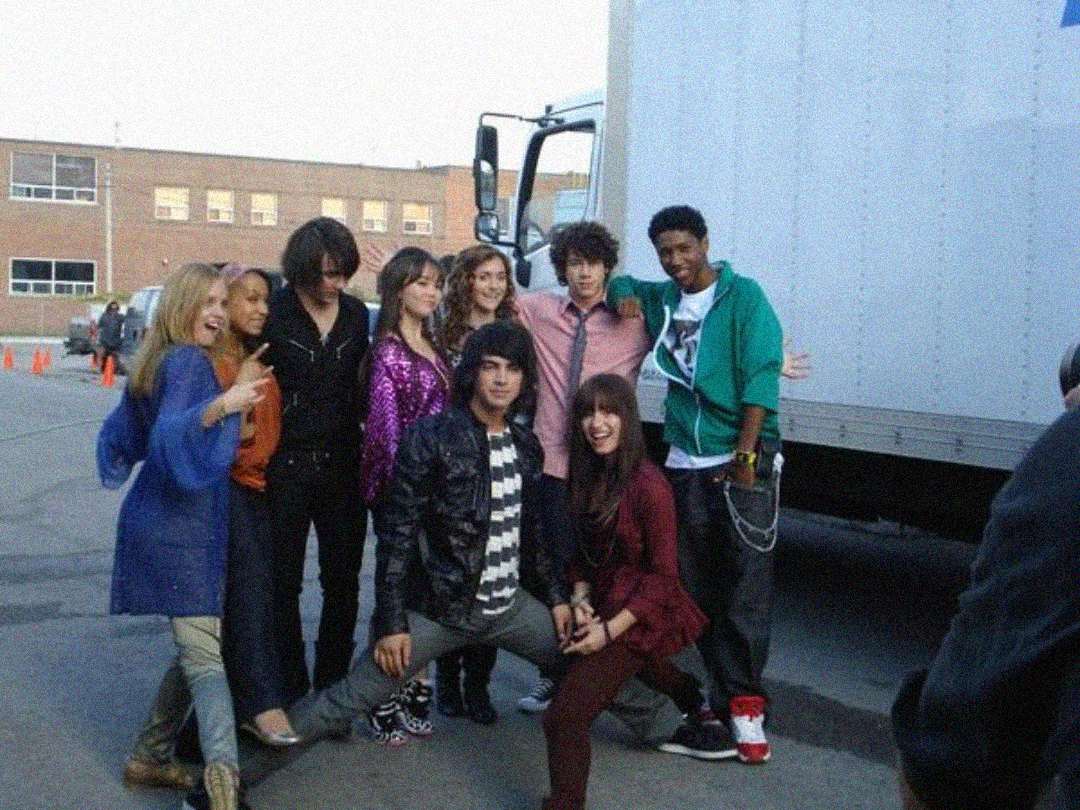 A group of eight young people pose together and smile outside near a large white truck and brick building. Most are standing close, one person kneels in front, and another stands with arms outstretched.