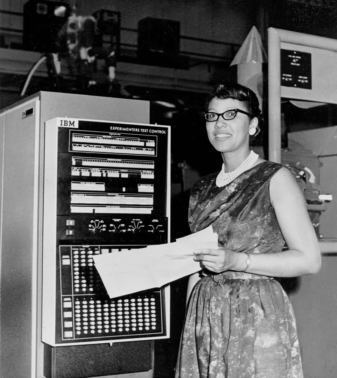 A woman in a patterned dress and glasses holds papers while standing next to an IBM Experimenter’s Test Control computer, featuring switches, dials, and indicator lights, in a mid-20th-century lab setting.
