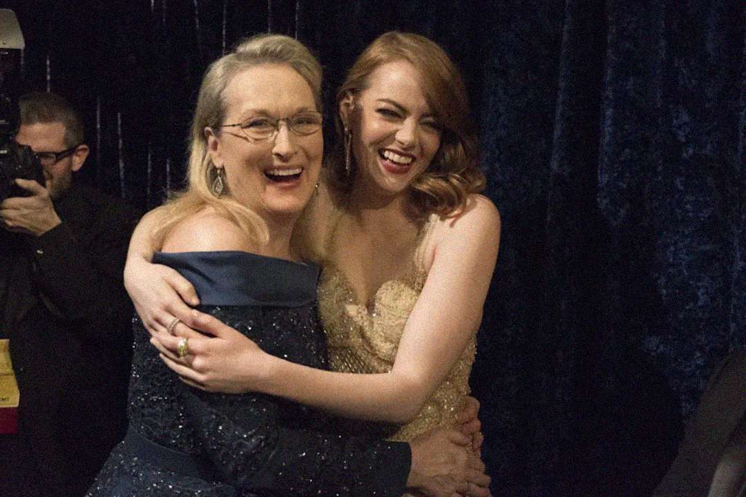 Two women in formal dresses smile and embrace while posing for a photo at an event. One wears glasses and a navy gown, the other has red hair and a strapless, sparkling dress. A photographer stands in the background.