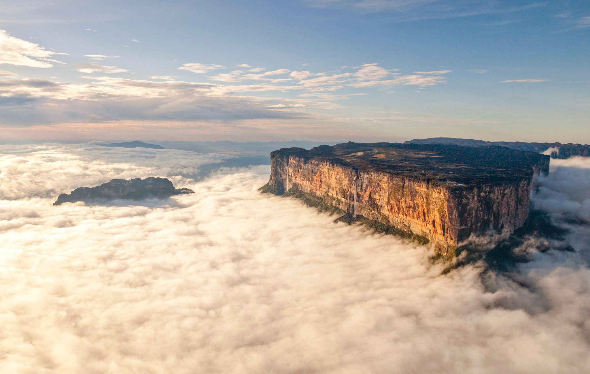 A massive, flat-topped mountain rises above a sea of clouds during sunrise or sunset. The sunlight casts a warm glow on the cliff faces, while the sky above is clear with scattered clouds on the horizon.