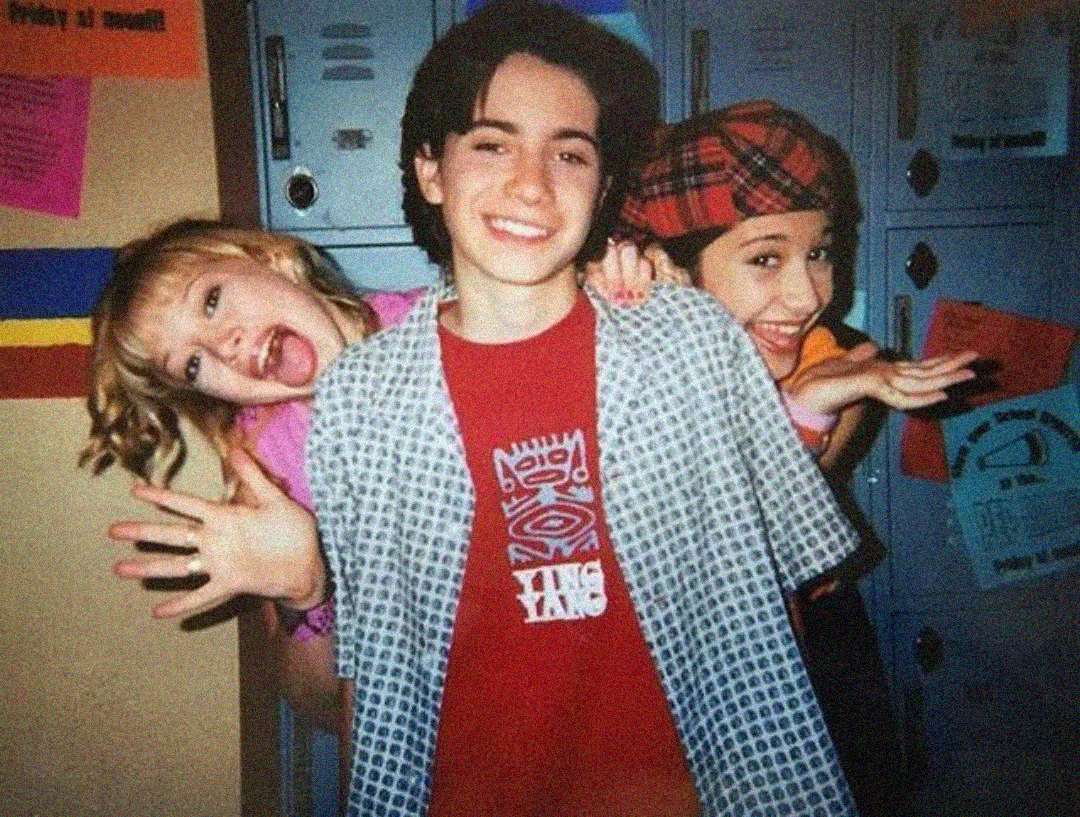 Three smiling kids pose in front of blue lockers, with one boy in the center wearing a red shirt and patterned shirt, and two girls, one on each side, making playful faces and gestures toward the camera.