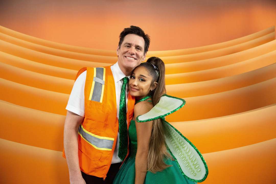 A man in a safety vest and a woman in a green fairy costume with wings stand closely together, smiling, in front of an orange, wavy background.