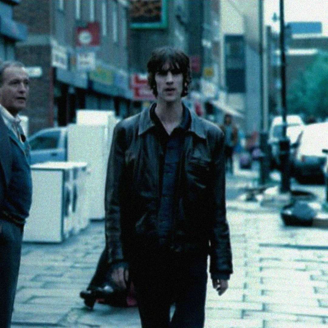 A man in a dark leather jacket walks down a wet city sidewalk, looking ahead. Another man stands to the side, and parked cars and storefronts line the street in the background.
