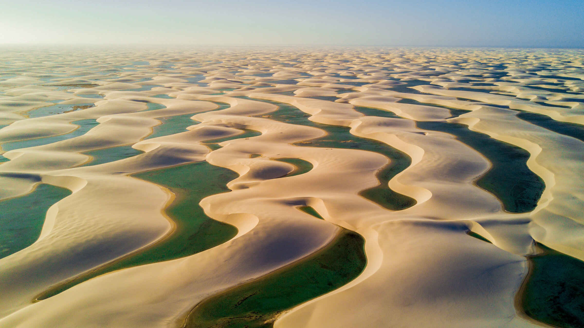 Aerial view of a vast desert landscape with rolling sand dunes and scattered blue-green lagoons between them under a clear sky.