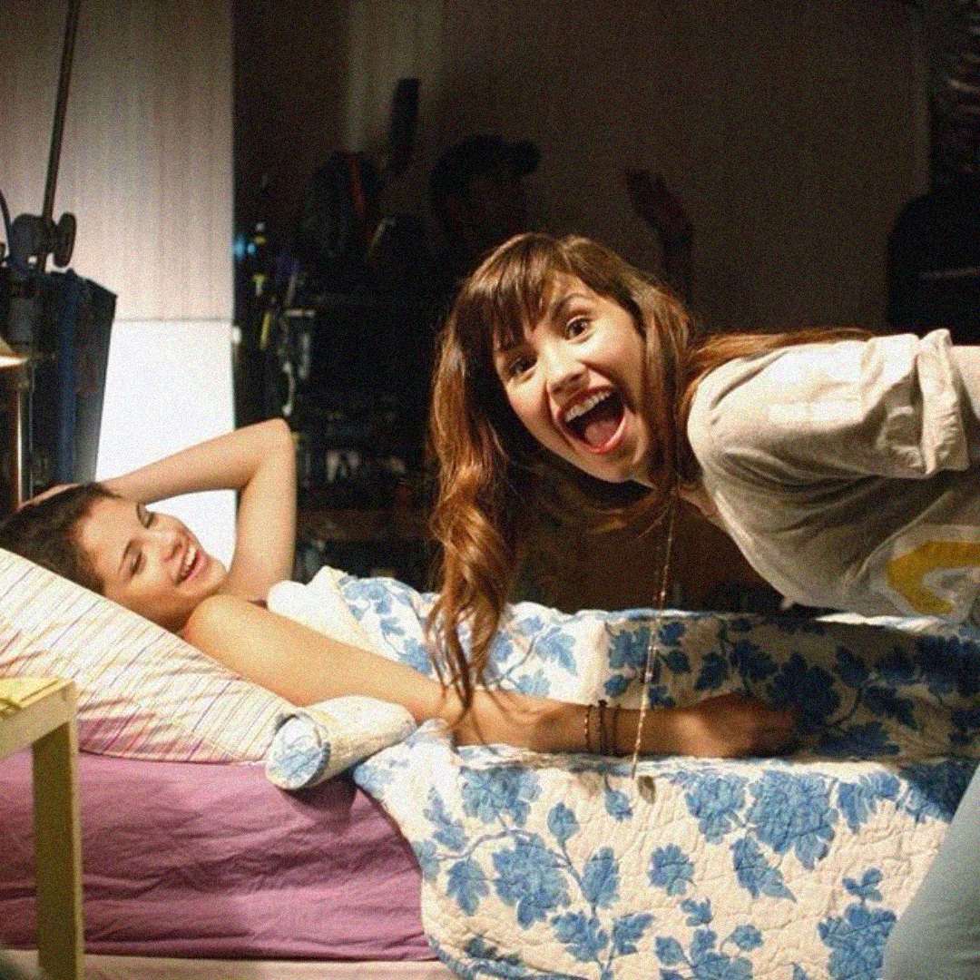 Two young women are in a cozy bedroom. One is lying in bed smiling, while the other leans over her, grinning playfully at the camera. The mood is cheerful and lighthearted.