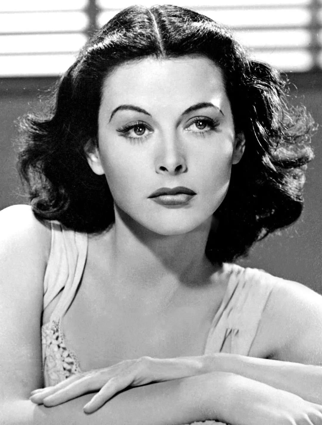 Black and white portrait of a woman with wavy dark hair, arched eyebrows, and light lipstick, resting her arms on a surface and gazing slightly to the side. Horizontal blinds appear in the background.