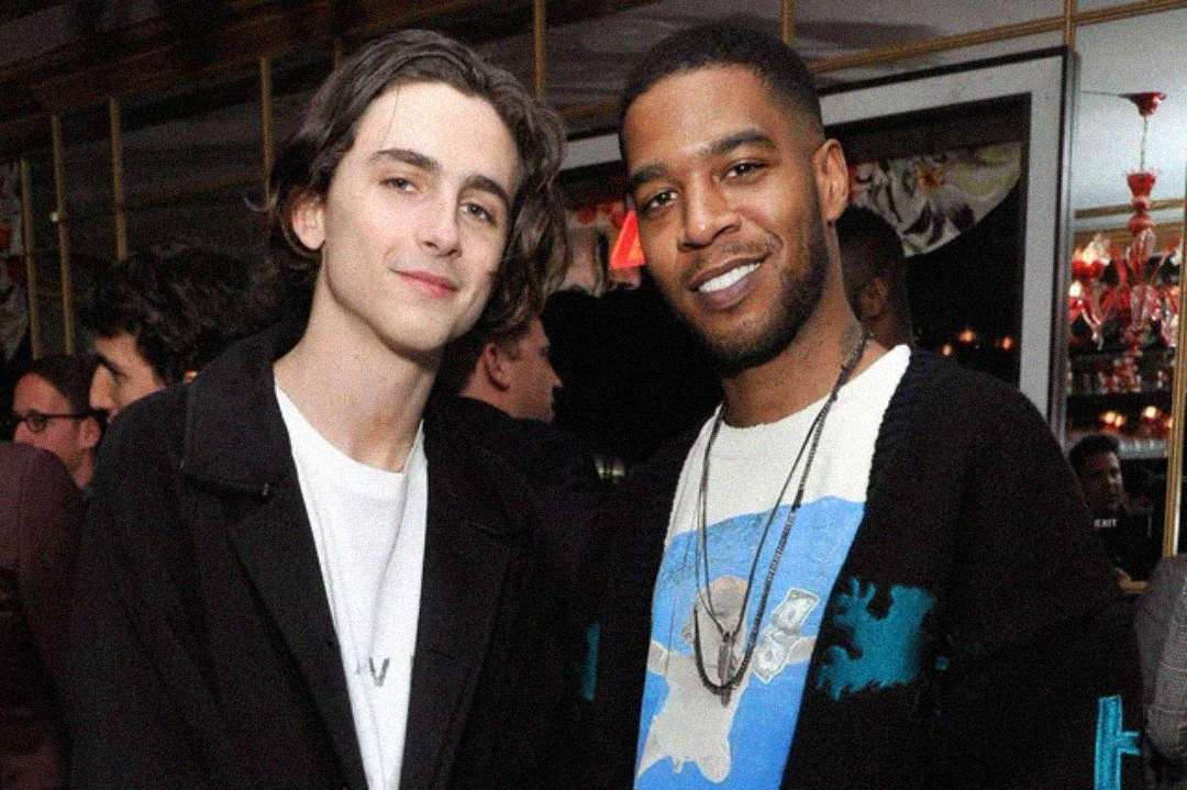 Two men are standing close together at an indoor social event, smiling at the camera. One has wavy brown hair and is wearing a white shirt and dark jacket, while the other wears a necklace and a patterned sweater.