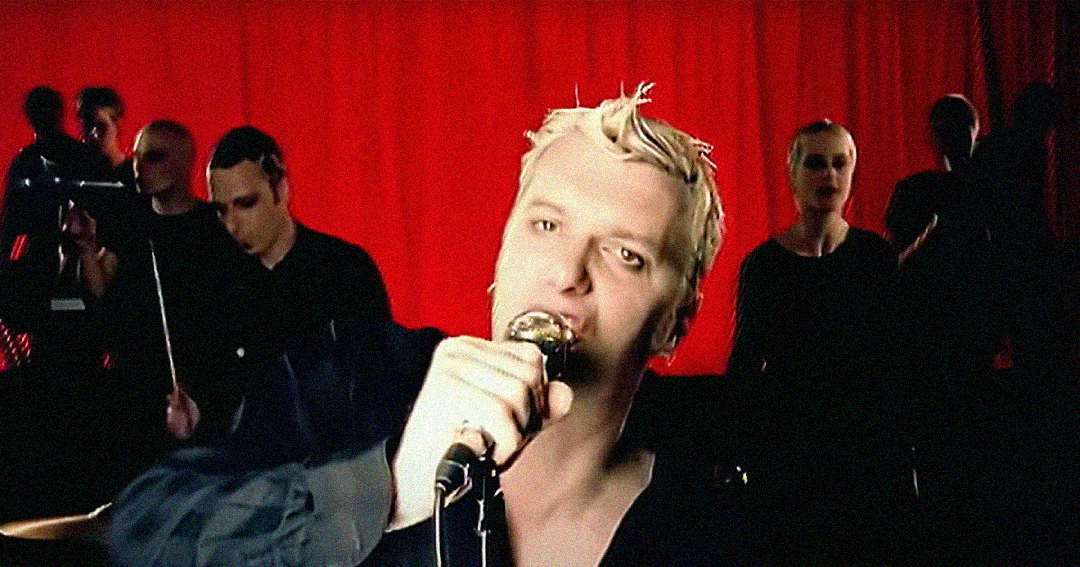 A person with short, spiky blond hair sings into a microphone in front of a bright red curtain, with band members playing instruments in the background.