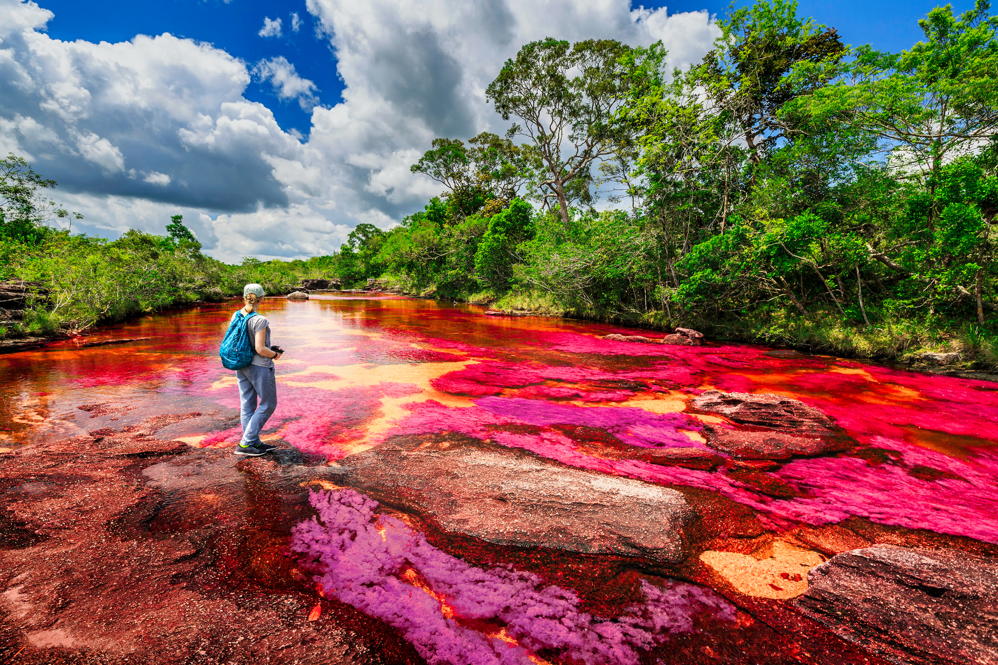 A person with a blue backpack stands on colorful, pink-red rocks by a vibrant river surrounded by lush green trees under a partly cloudy sky. The river has striking hues of red, orange, and purple.