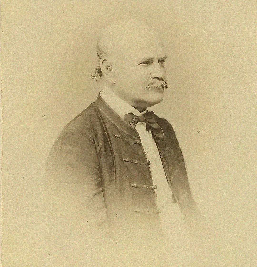 Sepia-toned portrait of an older man with a mustache, balding head, and side hair, wearing a traditional dark jacket and bow tie, facing slightly to the right.