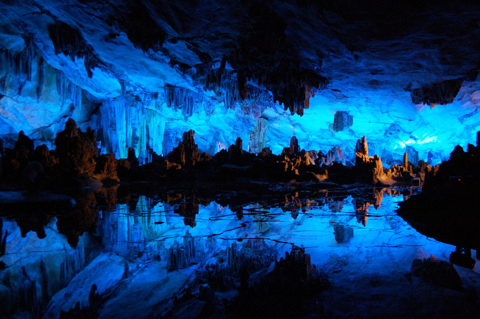 An underground cave filled with stalactites and stalagmites is brightly illuminated with blue lights, creating reflections on a still pool of water on the cave floor.