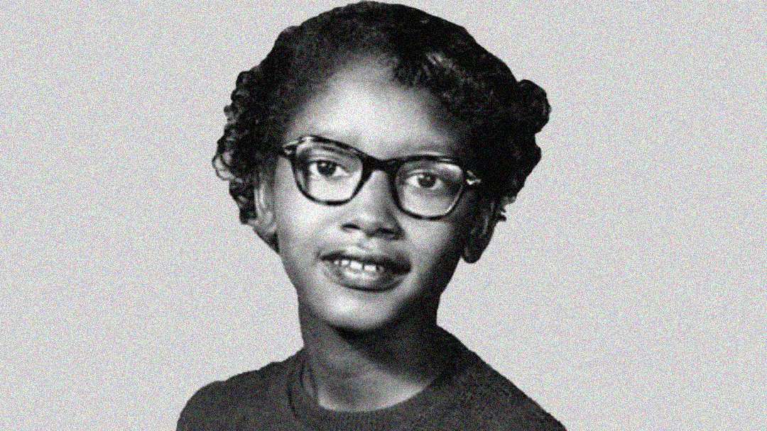 Black and white portrait of a young Black girl with short curly hair, wearing glasses and a dark top, smiling slightly and looking towards the camera against a plain background.