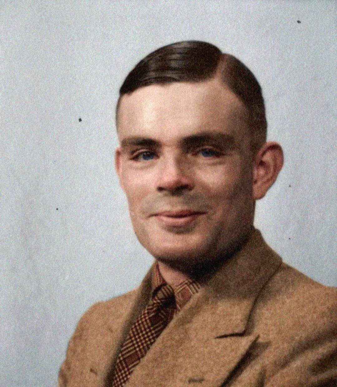A man with short, neatly combed brown hair wears a light brown suit jacket, checked tie, and tan shirt, smiling slightly in front of a plain light background.