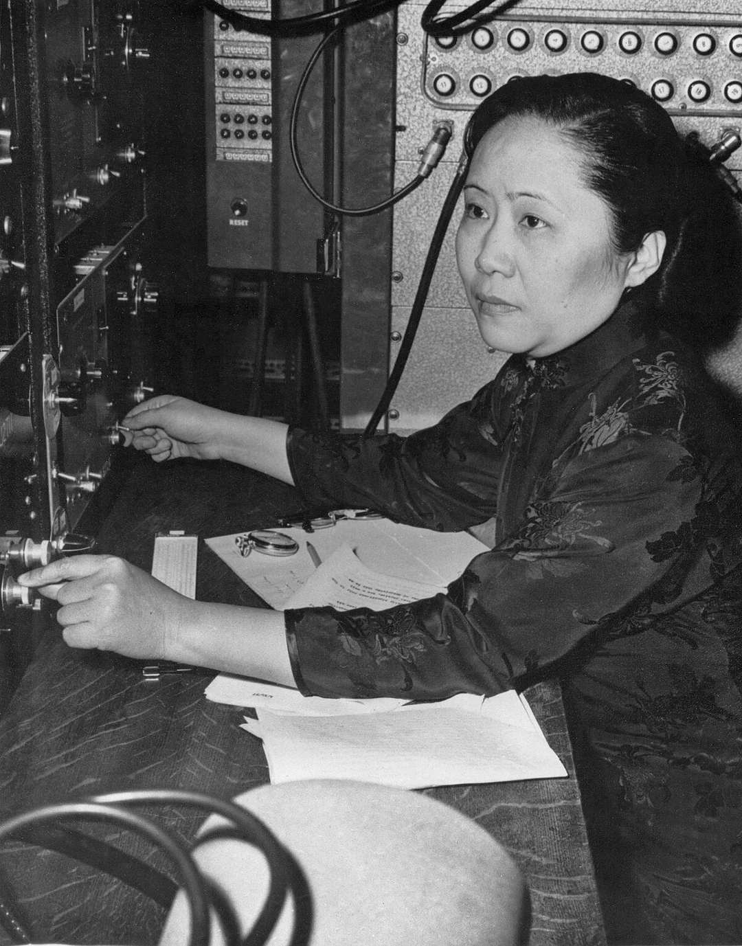 A woman in a patterned dress sits at a control panel, adjusting dials and switches. Papers and a pen lie on the table in front of her. Electronic equipment and cables fill the background.