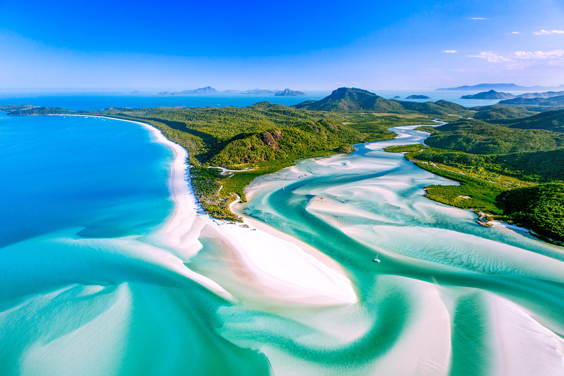 Aerial view of a tropical coastline with turquoise water, white sandy beaches, lush green hills, and winding tidal inlets under a bright blue sky.