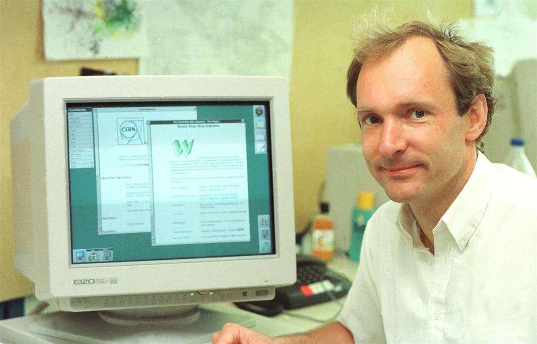 A man with light hair and a white shirt sits next to an old computer monitor displaying an early web browser interface, with text and graphics visible on the screen.