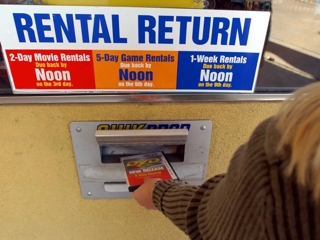 A person returns a DVD to a rental return slot below a sign detailing return times for 2-day movie rentals, 5-day game rentals, and 1-week rentals, all due back by noon on specified days.