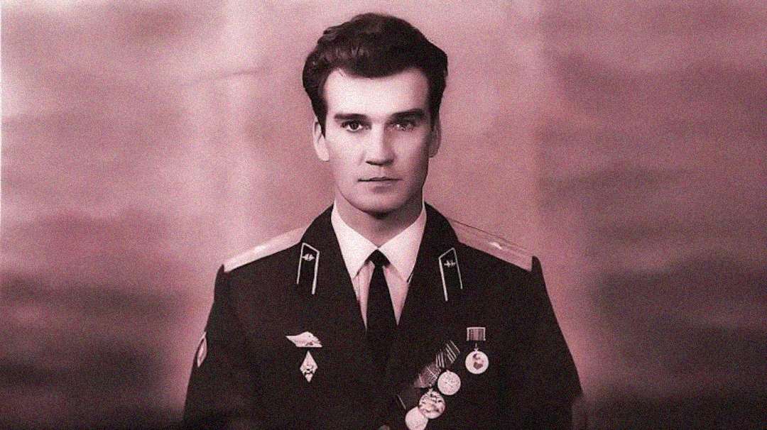 A young man in a military uniform with several badges and medals on his chest faces the camera with a neutral expression. The image is in sepia tones, giving it a vintage appearance.