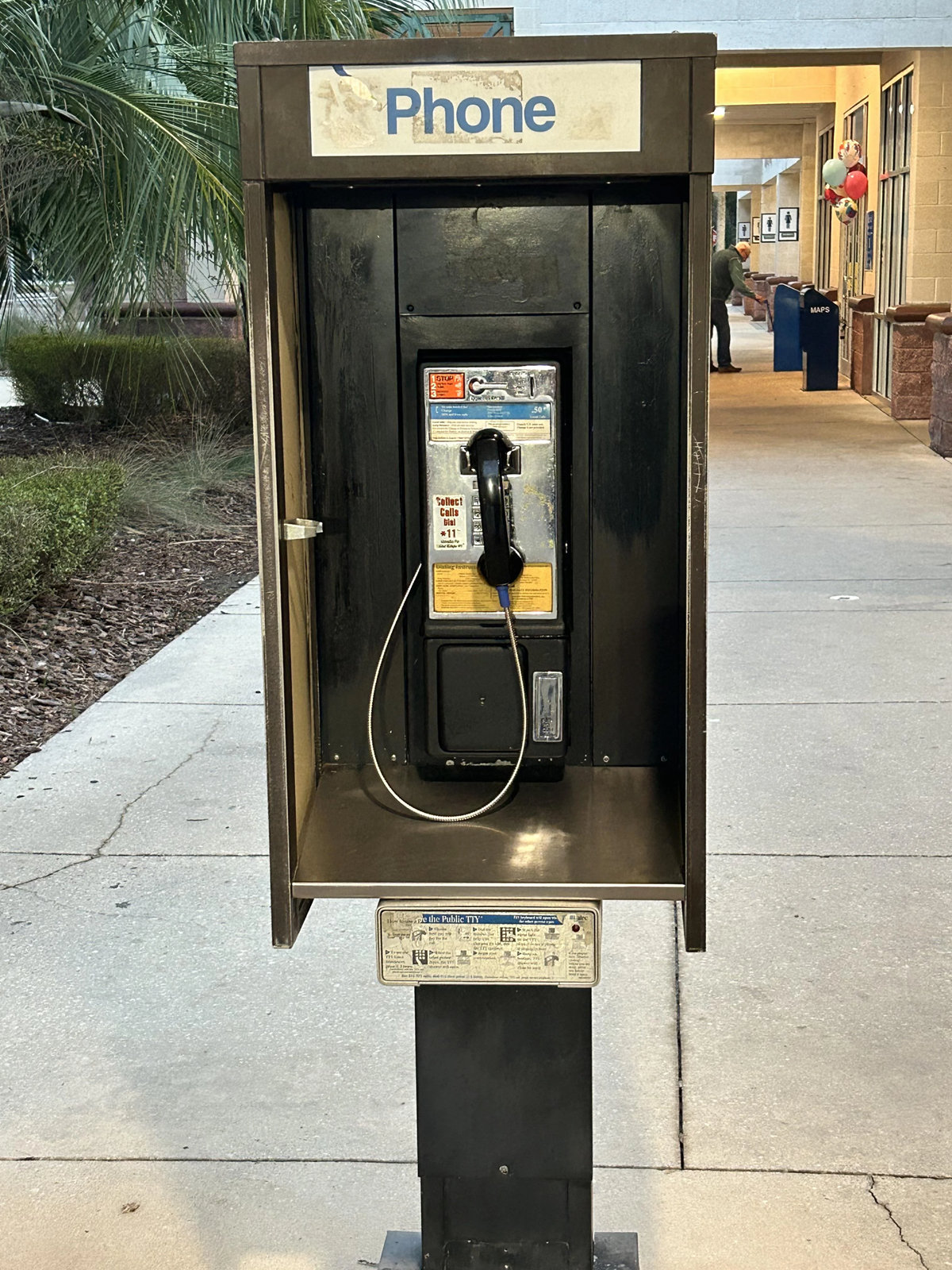 A public payphone booth with a handset and coin slot stands on a sidewalk near a building entrance, with palm trees and people visible in the background.