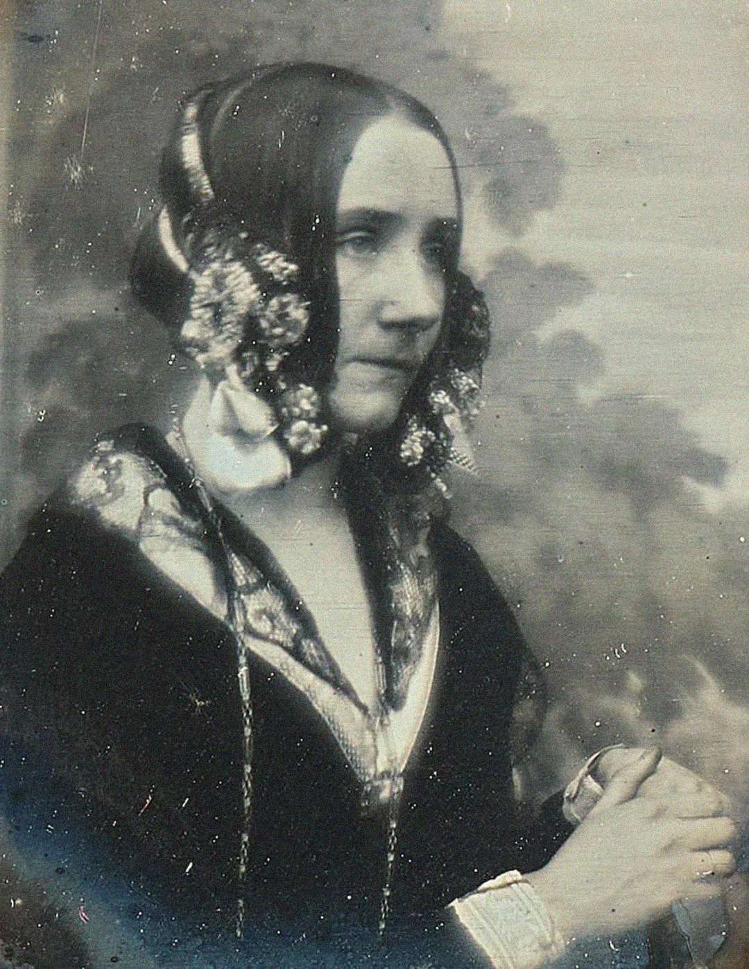 Black and white portrait of a woman in Victorian-era clothing, with elaborate hair accessories, looking slightly to the side. She holds something in her hands and has a calm, composed expression.