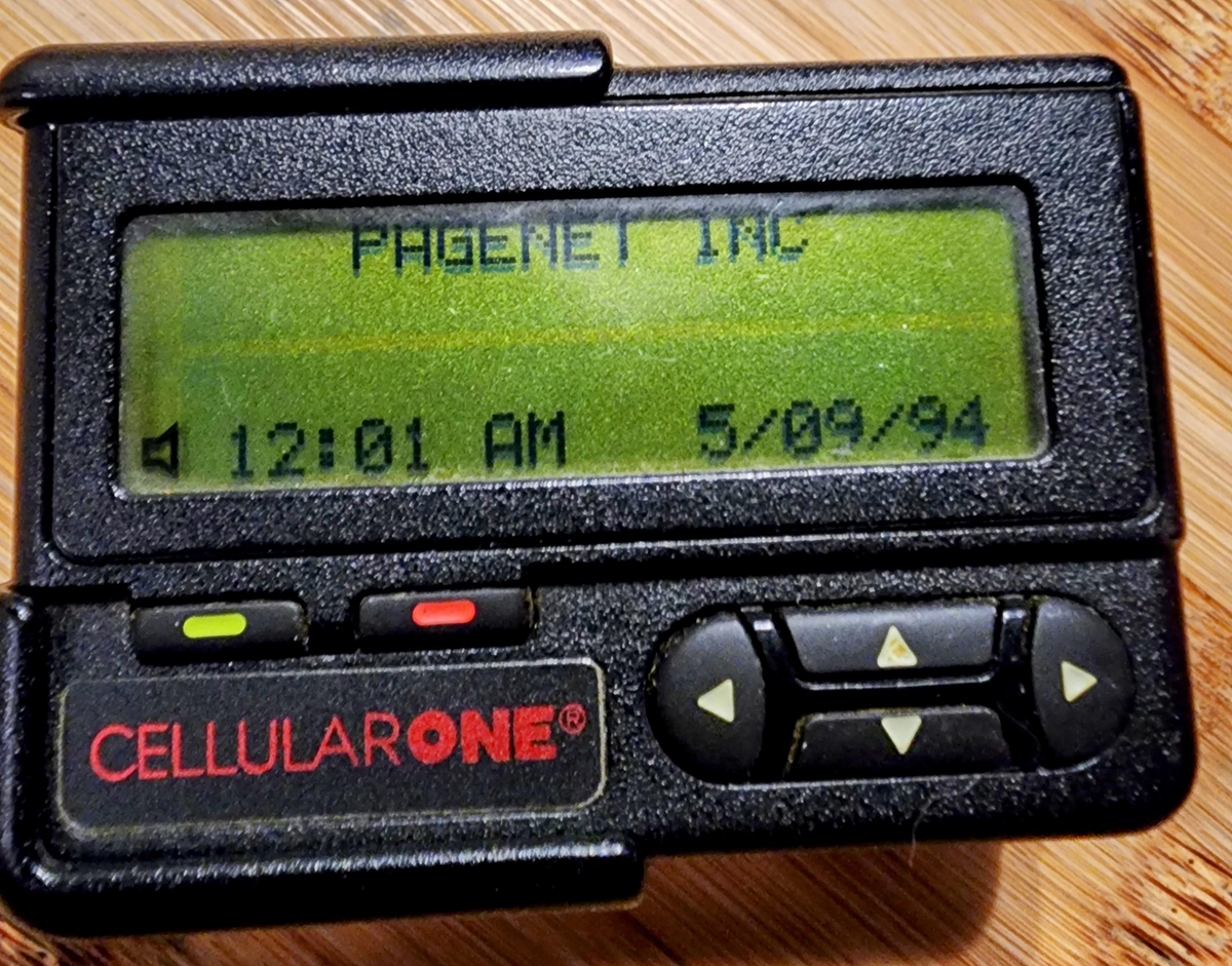 A close-up of a black pager displaying "PAGERET INC," the time "12:01 AM," and the date "5/09/94" on its green screen. The pager has buttons and a red "CELLULARONE" logo on the lower left.