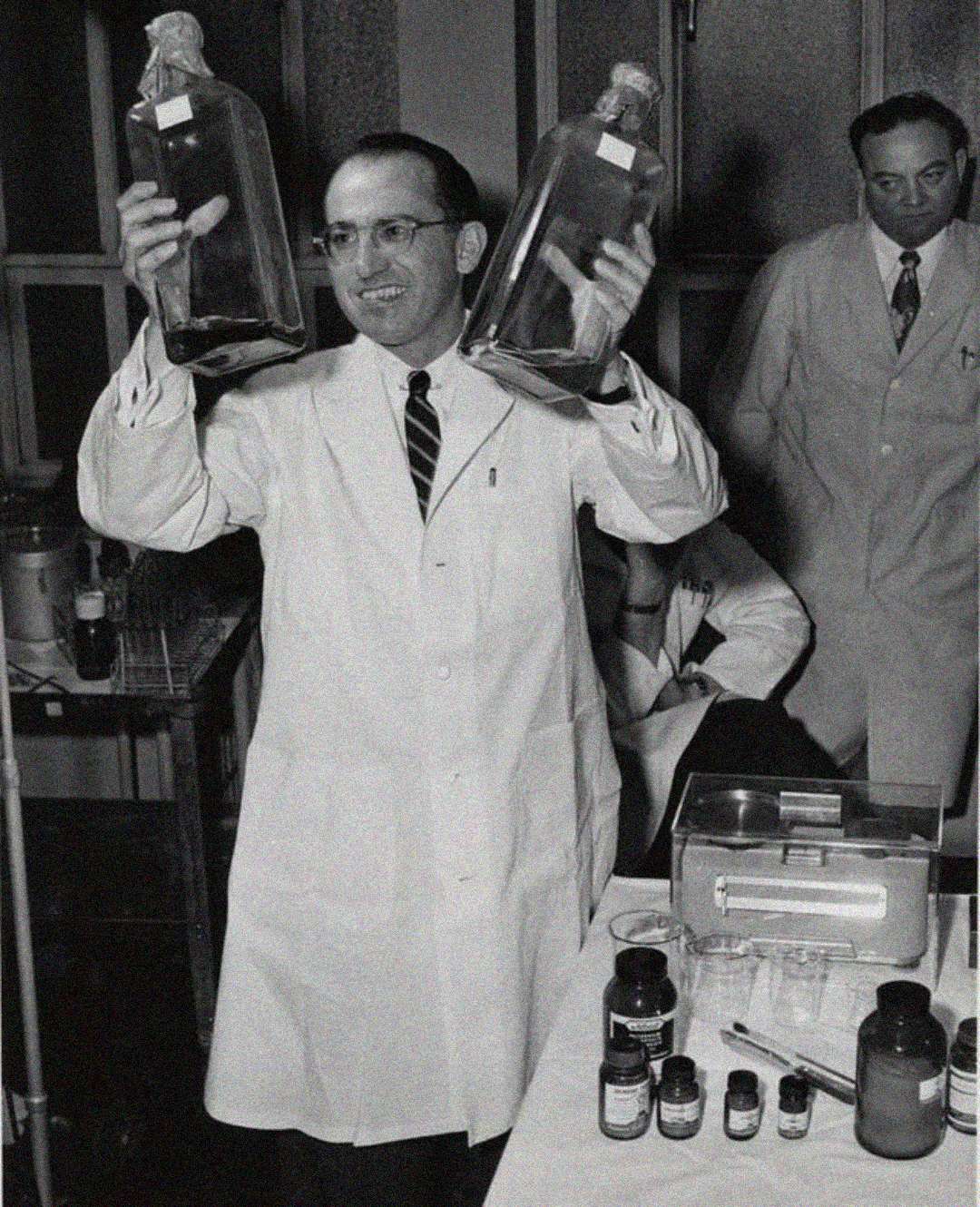 A man in a white lab coat holds up two large glass bottles, smiling. Another man in a lab coat stands behind him. Laboratory equipment and bottles are visible on a table nearby.