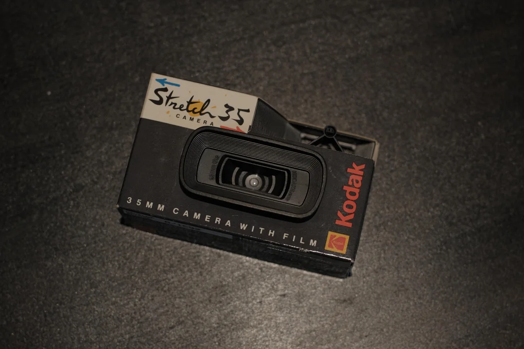 A vintage Kodak Stretch 35 camera with a black body and retro branding lies on a dark textured surface. The camera features "35mm camera with film" text and a prominent circular lens.