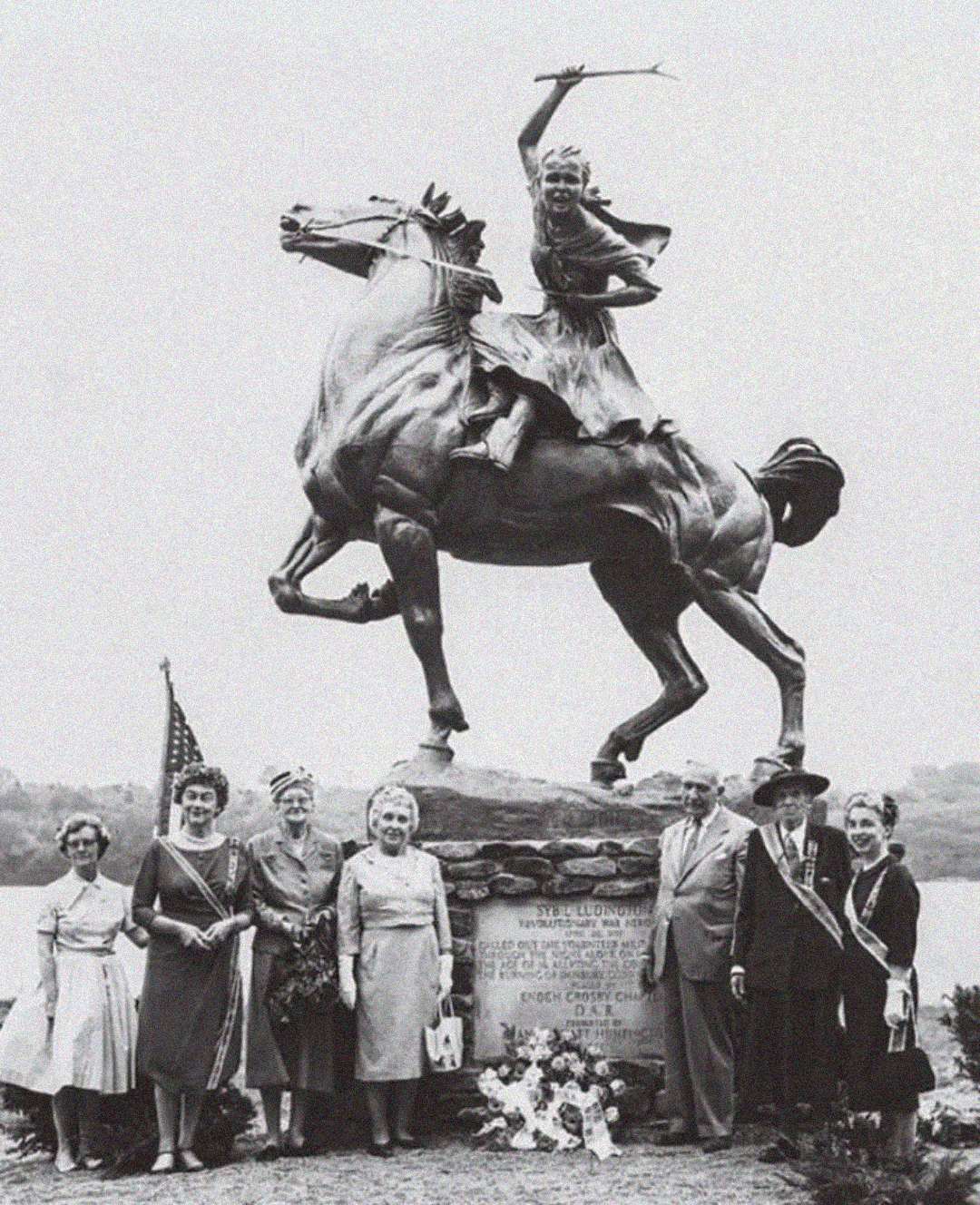 A black-and-white photo shows seven people standing in front of a large equestrian statue of a woman on a rearing horse. Some are wearing formal clothes and sashes, with an American flag and flowers at the statue's base.