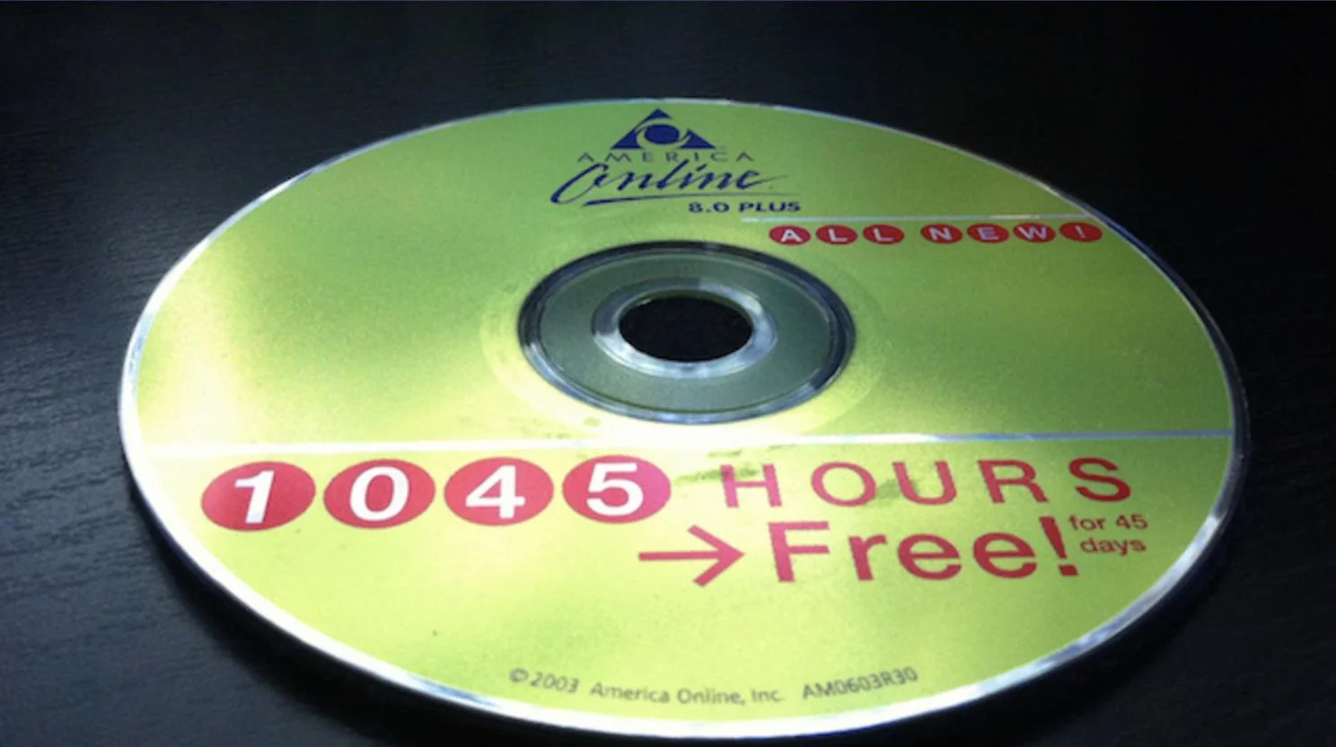 A yellow America Online 8.0 Plus CD offers "1045 hours free for 45 days," featuring red and white text and the AOL logo, lying on a dark surface.