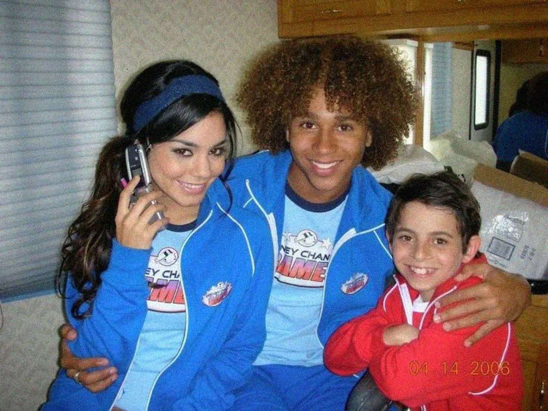 Three smiling young people in blue and red Disney Channel Games outfits sit close together indoors. One girl holds a phone, another boy is in the middle, and a younger boy sits beside them. Date stamp reads "04 14 2006.