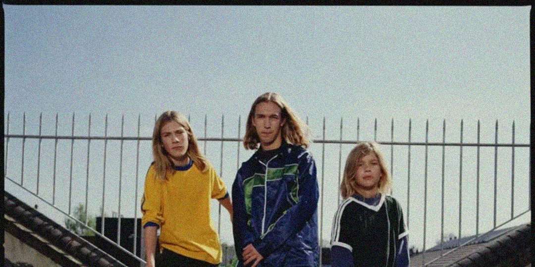Three young people with long hair stand outdoors in front of a metal fence, wearing casual sportswear tops in yellow, blue, and black. They look directly at the camera under a clear sky.