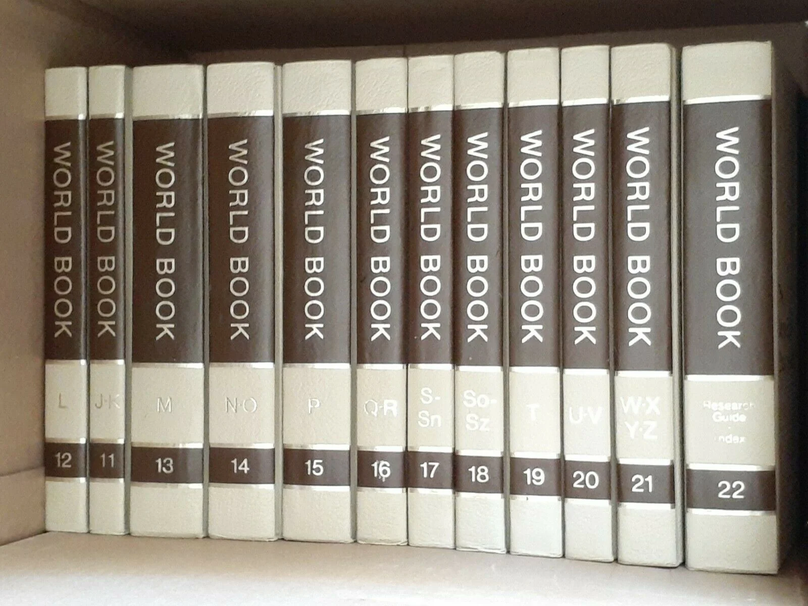 A shelf with several volumes of the World Book Encyclopedia, arranged from L to Z with their corresponding volume numbers 12 to 22. The books have white and brown spines with bold titles.