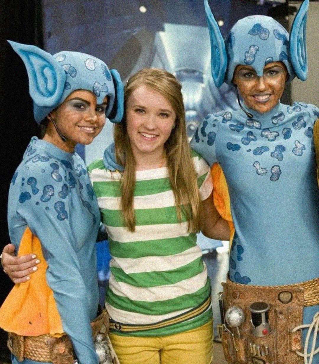 Three people pose together. The person in the center wears a green and white striped shirt and yellow pants. The two on either side wear blue mouse costumes with large ears, face paint, and utility belts. All are smiling.