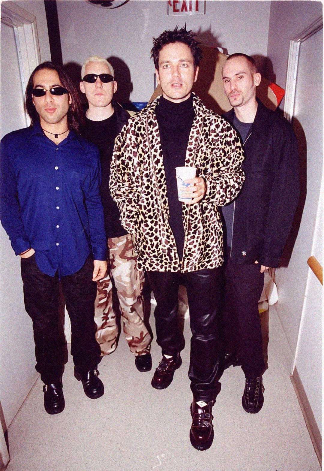 Four men stand in a hallway. The man in front wears a leopard print jacket and holds a cup, while the others wear dark shirts and sunglasses. One has bleach-blond hair and camouflage pants. They all pose for the camera.
