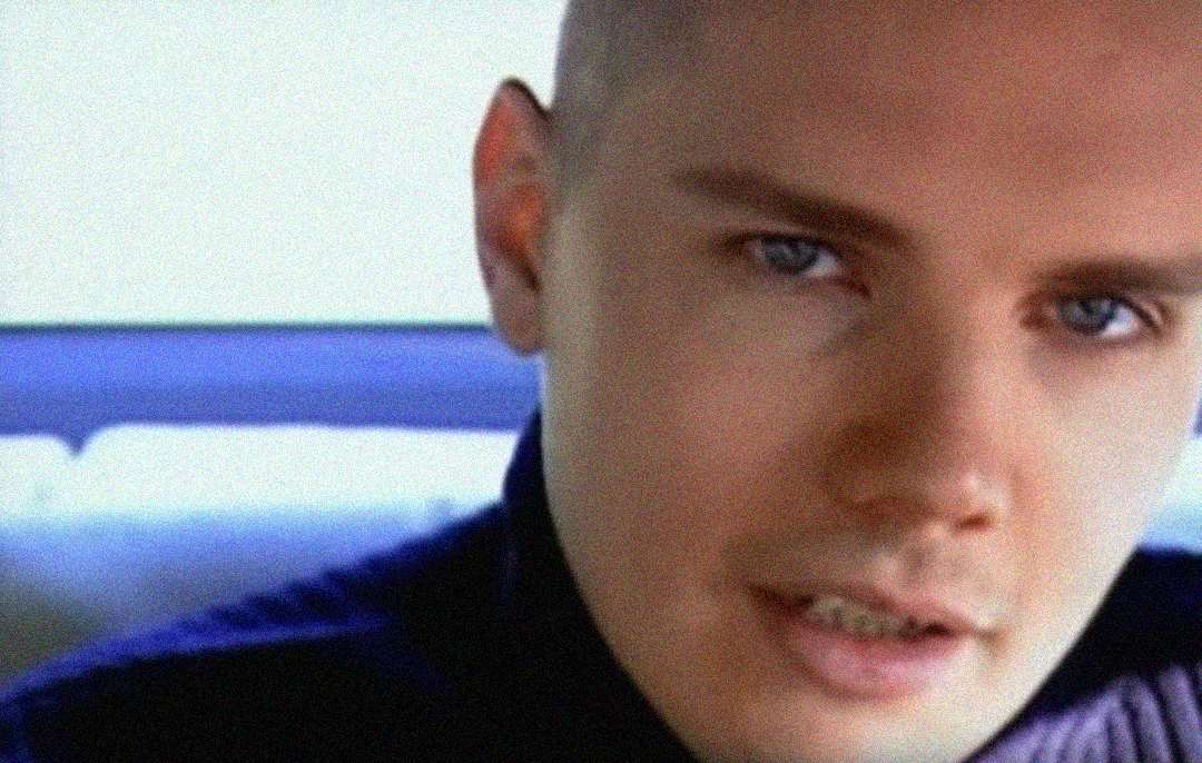 A close-up of a person with light skin, blue eyes, and a shaved head, sitting in a car. The background is out of focus, highlighting the person's face and expression.