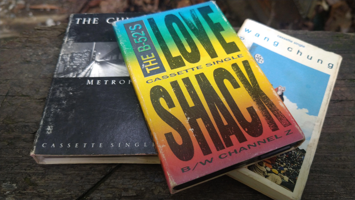 Three vintage cassette singles are stacked on a wooden surface: "The B-52's Love Shack," "The Church Metropolis," and "Wang Chung Dance Hall Days." The covers are colorful and slightly worn.