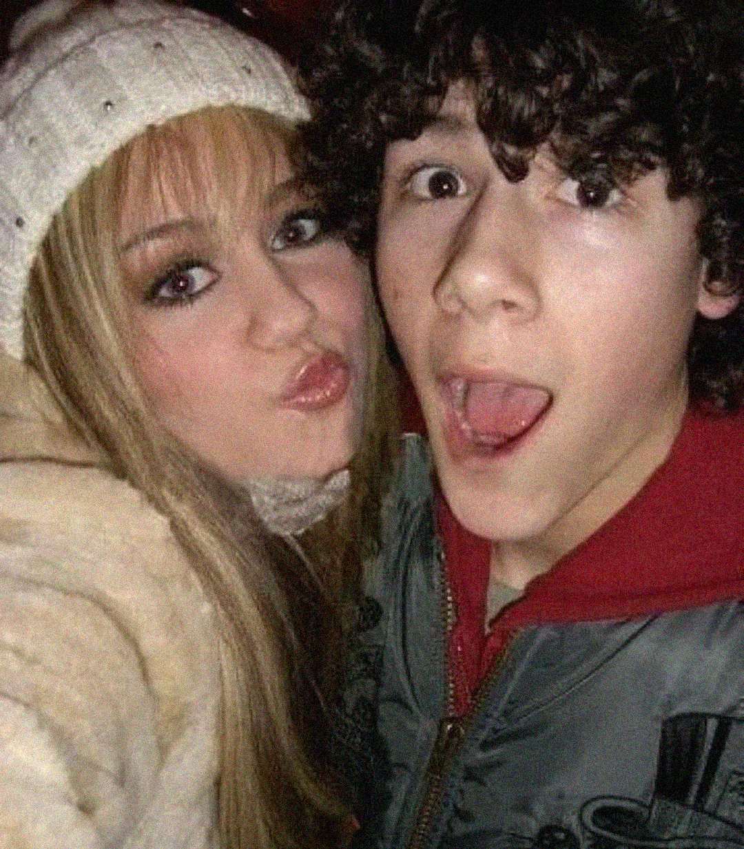 Two teenagers posing for a selfie; the girl in a white beanie and furry coat is pouting, while the boy beside her has curly dark hair and an open-mouthed surprised expression.