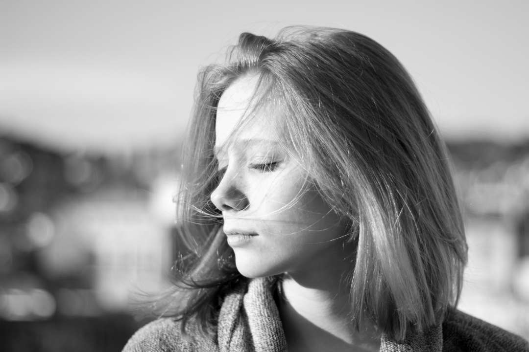 Black and white portrait of a young woman with light hair, eyes closed, and a calm expression. She is facing slightly to the left with the wind gently blowing her hair. The background is blurred.