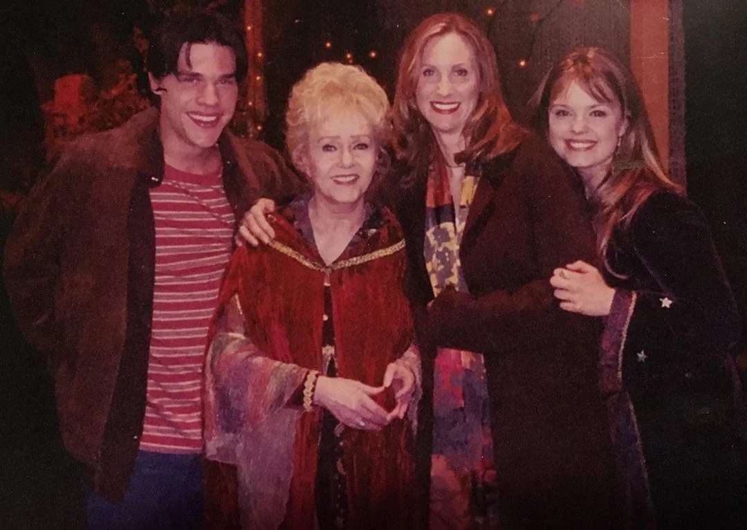 Four people stand close together, smiling at the camera. One older woman in the center wears a red shawl, while the three younger adults beside her are dressed warmly in coats. They appear to be in a festive or magical setting.