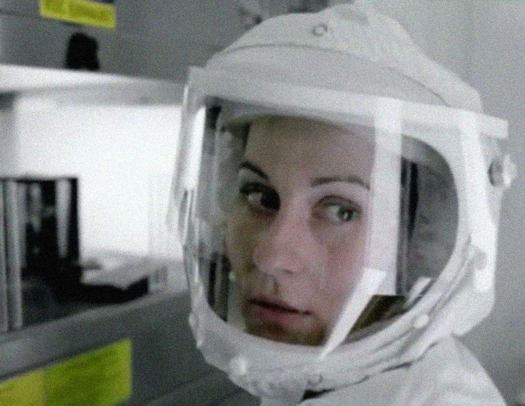 A person wearing a white hazmat suit and a clear face shield looks toward the camera, standing in a laboratory setting with shelves and equipment in the blurred background.