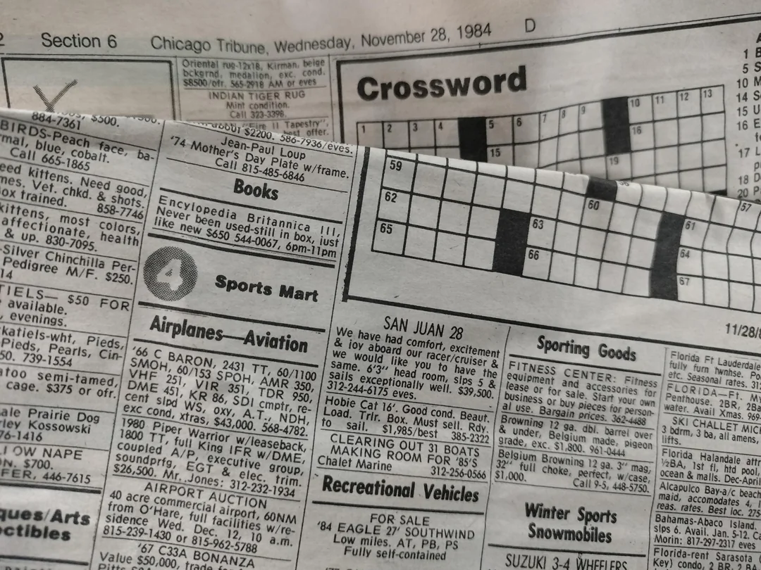 A close-up of a vintage newspaper page from the Chicago Tribune dated November 28, 1984, showing a crossword puzzle and classified ads sections for books, sports, airplanes, and recreational vehicles.