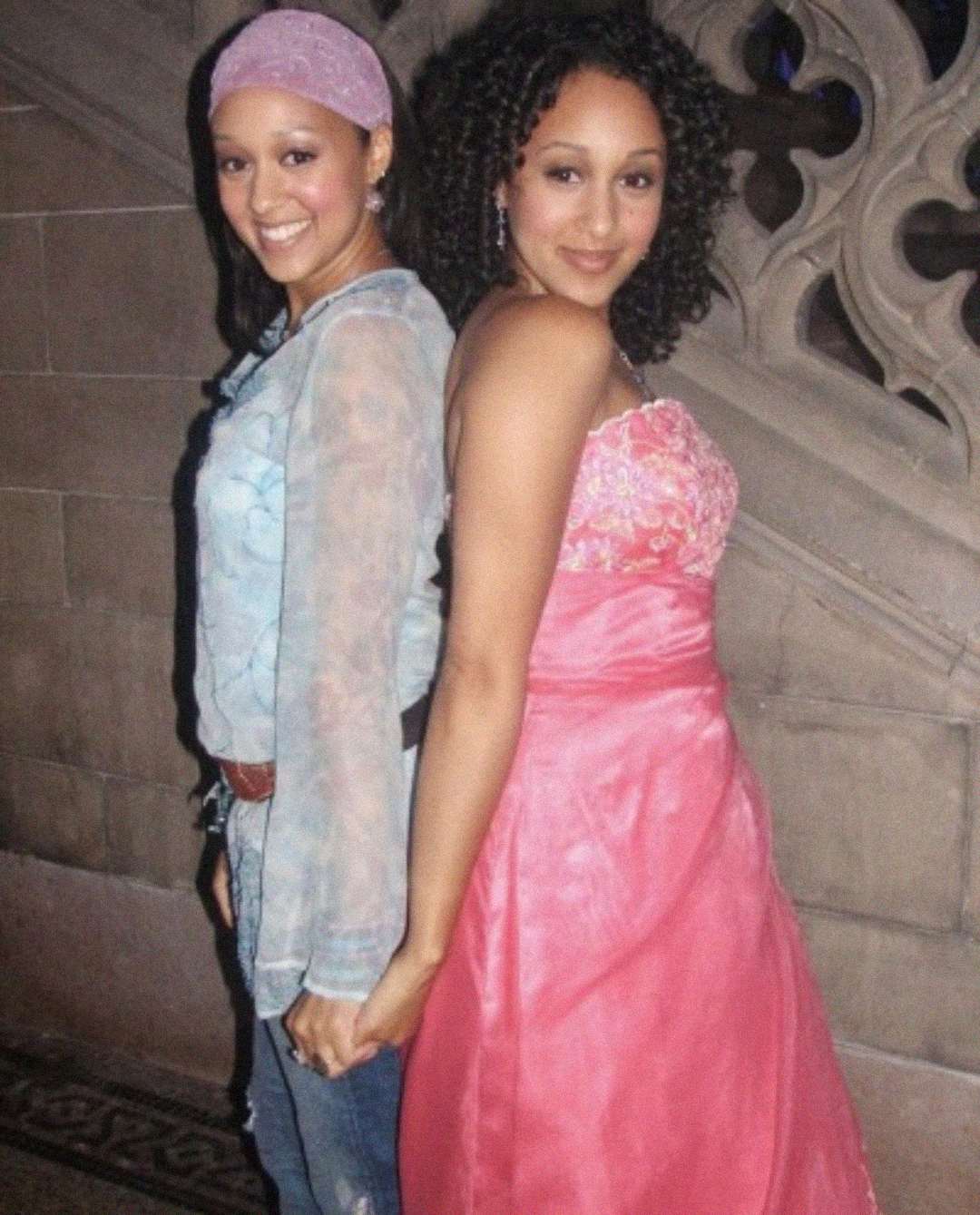 Two women stand back to back holding hands. One wears a casual outfit with jeans, a pink headband, and a sheer blue top. The other wears a pink strapless gown. They both smile and pose in front of a carved stone wall.