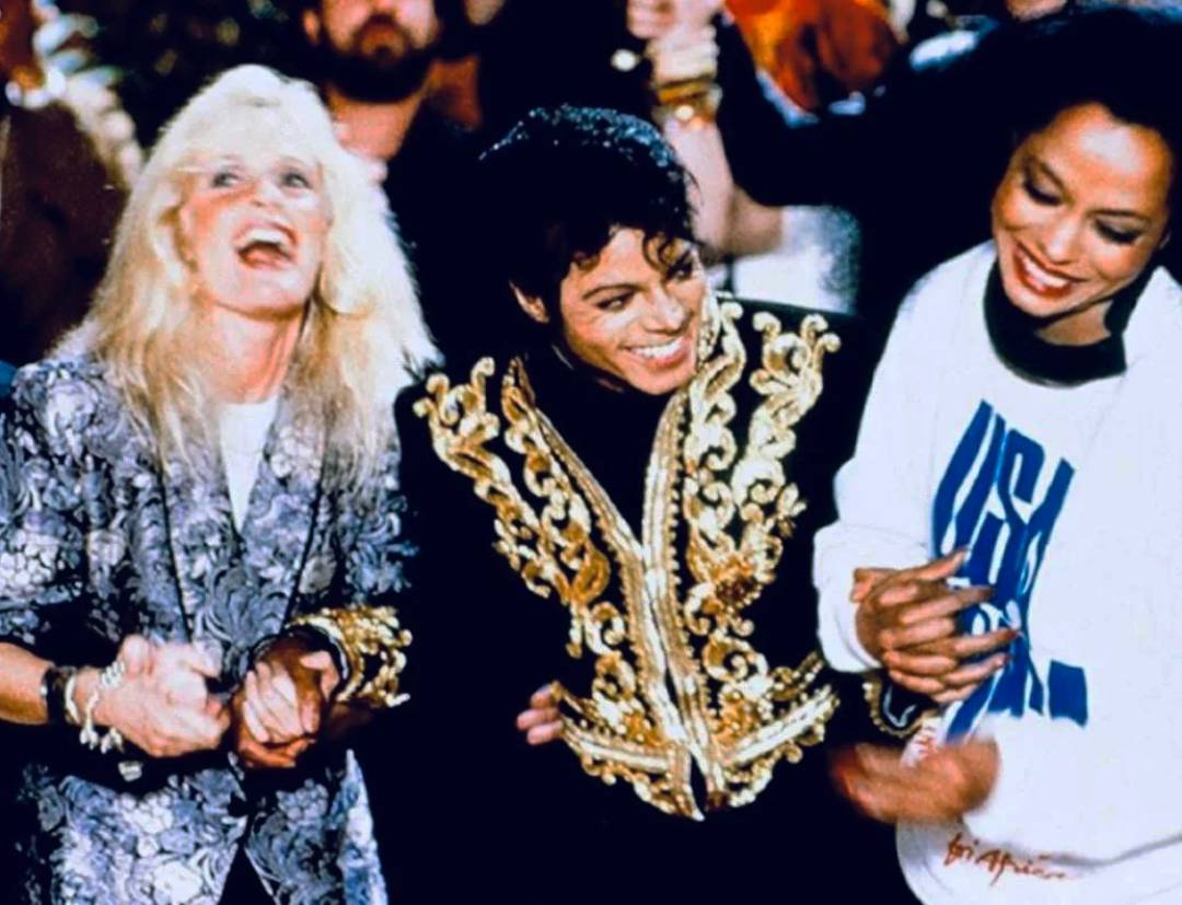 Three people laugh and hold hands at an event. The person in the center wears a black and gold ornate jacket, while those on either side wear patterned jackets and a white sweatshirt. The atmosphere is joyful and lively.