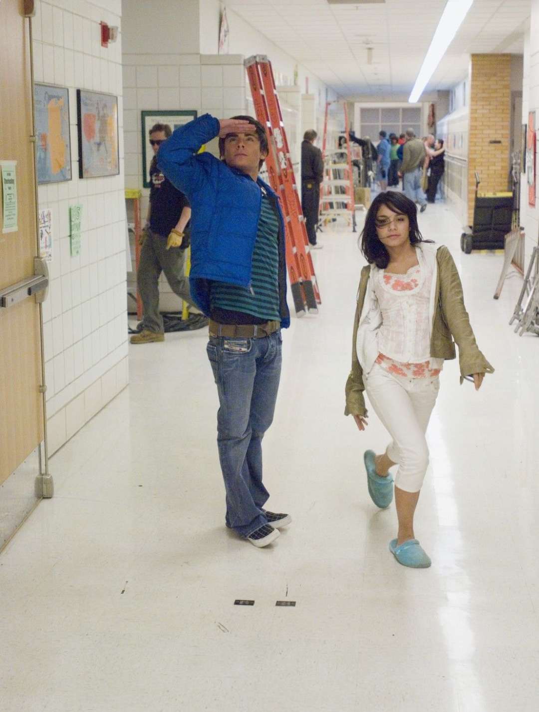 Two teenagers walk down a school hallway; the boy in a blue jacket looks up while the girl in a white outfit and blue slippers walks beside him. People, ladders, and equipment are seen further down the hallway.