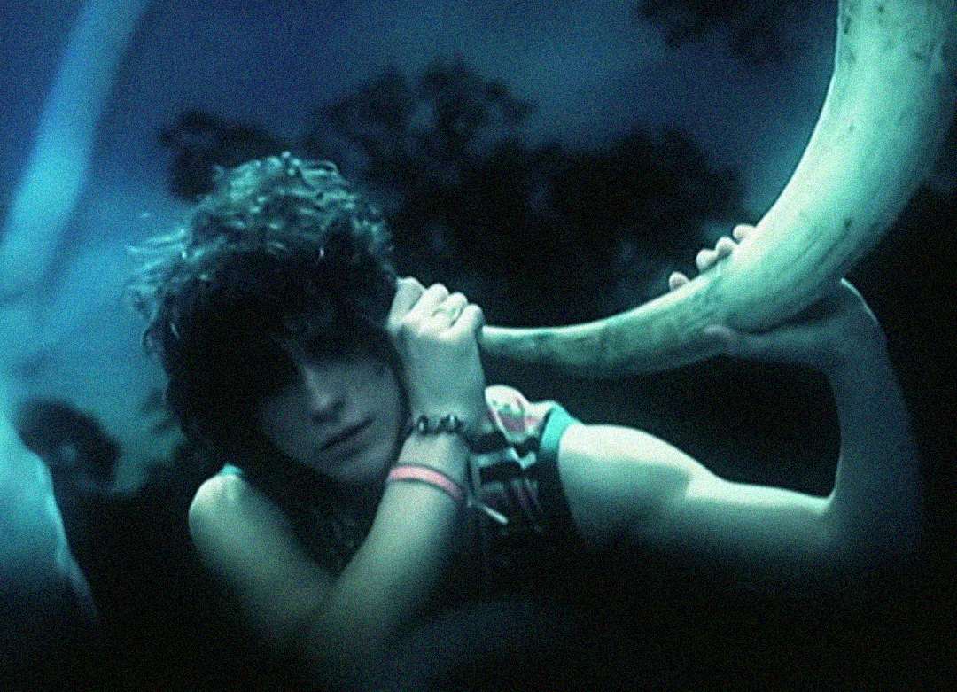 A person with curly hair holds a large curved horn to their ear, appearing to listen to it. The scene is dimly lit with a blue tint, giving it a mysterious, dreamy atmosphere.