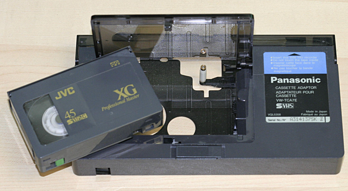 A JVC MiniDV videocassette is placed next to an open Panasonic VHS cassette adapter on a light wooden surface. The adapter has a label with model information visible on the side.