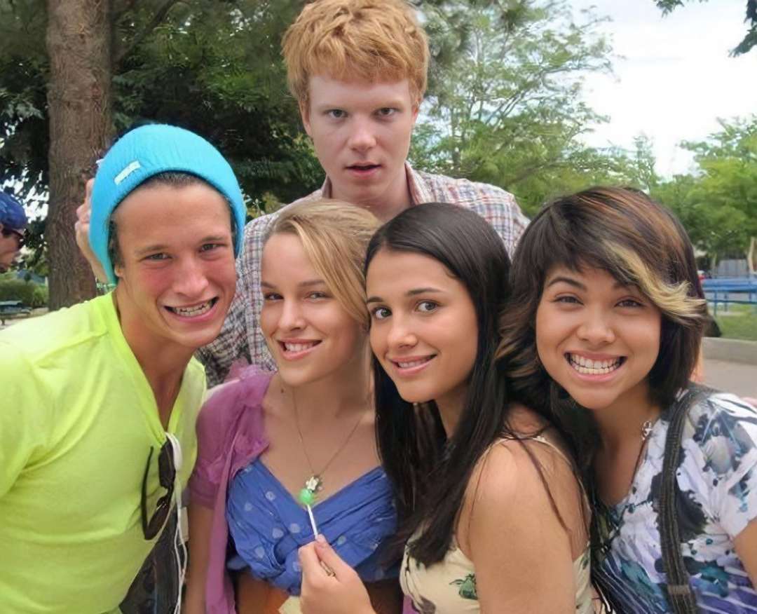 Five smiling young people pose closely together outdoors, with trees and greenery in the background. Four are standing in front, while one stands behind them, all appearing cheerful and happy.