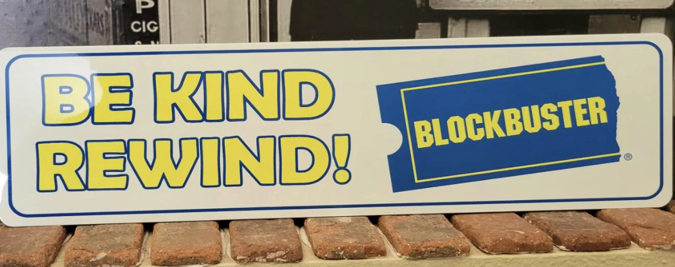 A rectangular sign with the text "BE KIND REWIND!" in yellow letters and the blue and yellow Blockbuster logo on a white background, resting on a brick surface.