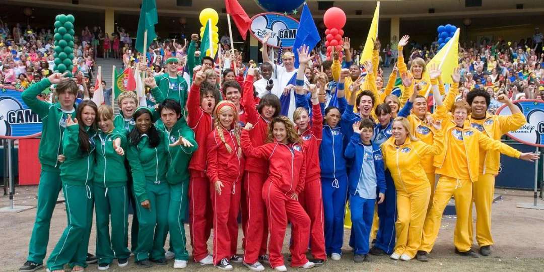 A large group of young people in red, green, blue, and yellow tracksuits stand together and smile for a group photo at an outdoor event, with cheering crowds and Disney Channel Game signs in the background.